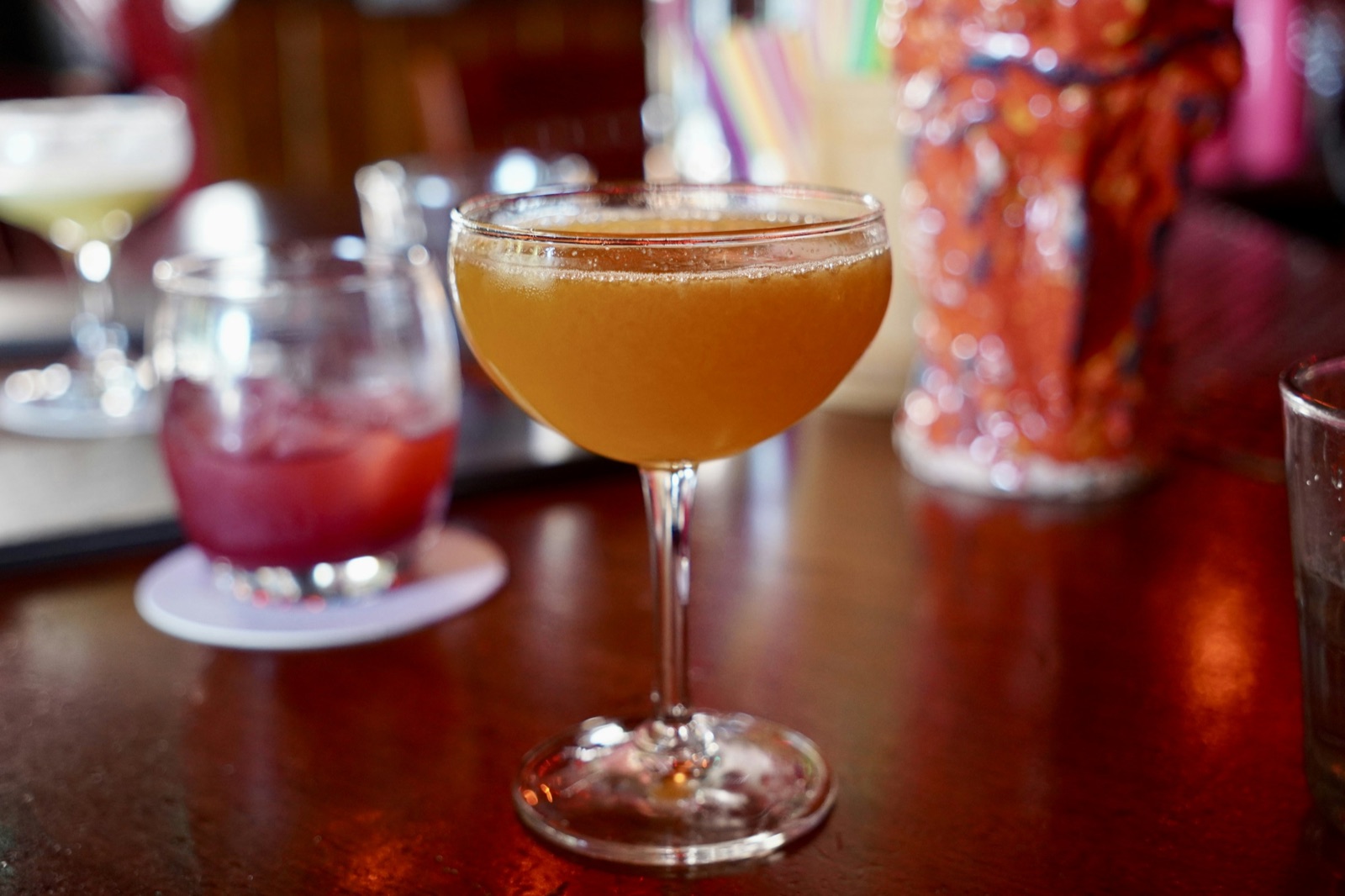 A golden cocktail in a coupe glass on a warmly lit bar